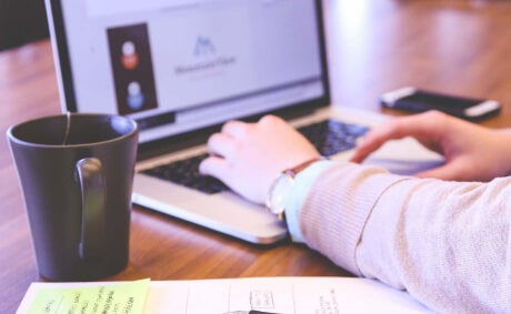 KICS_Digitale-Projektarbeit-und-Kundensupport Person arbeitet an einem Laptop mit geöffnetem Webbrowser, daneben Kaffeetasse und Notizen auf dem Schreibtisch.