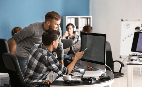 KICS_Moderne-Teamarbeit-und-agiles-Arbeiten Zwei Personen diskutieren vor einem großen Monitor in einem modernen Büro, weitere Teammitglieder arbeiten im Hintergrund.