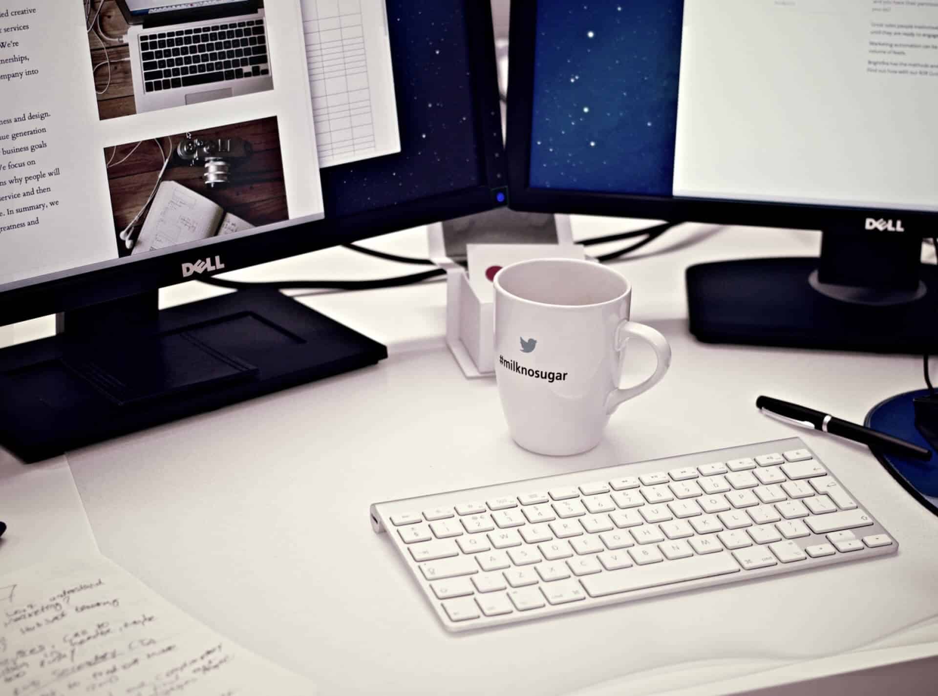 KICS_Moderner-Digitaler-Arbeitsplatz Aufgeräumter Schreibtisch mit zwei Monitoren, weißer Tastatur, Kaffeetasse und Notizen.