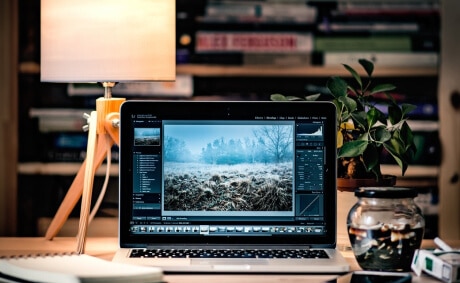 KICS_Modernes-Arbeitsumfeld-für-digitale-Services Laptop mit geöffneter Bildbearbeitungssoftware auf einem Schreibtisch mit Lampe, Pflanze und Büchern im Hintergrund.