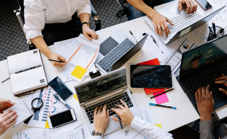 KICS_Offene-Zusammenarbeit-und-agiles-Teamwork Arbeitsgruppe mit Laptops, Tablets, Notizen und Dokumenten an einem gemeinsamen Tisch – Teamarbeit in Aktion.